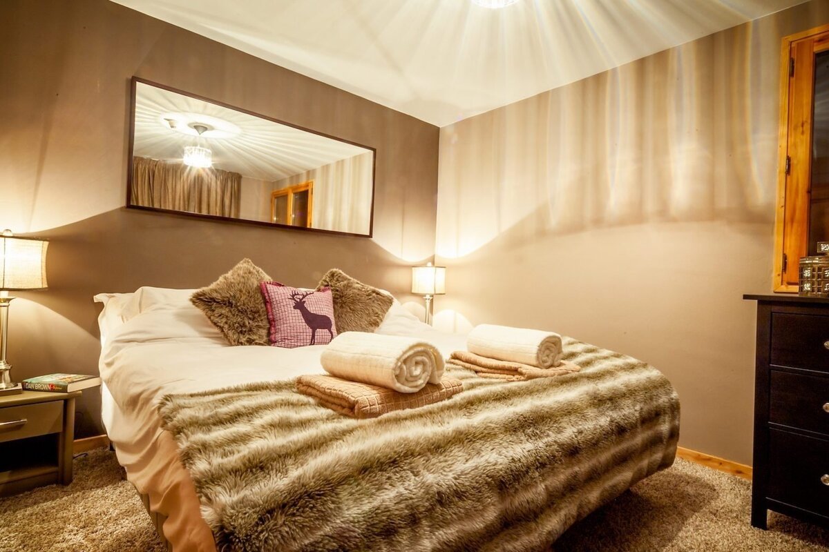 A cozy, warm bedroom features a large bed with a faux fur throw, deer-print pillow, and soft lighting. A mirror above the bed reflects the ceiling fixture.
