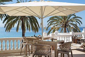 Sunny patio with wicker furniture under a white umbrella, overlooking a clear blue ocean and framed by palm trees.