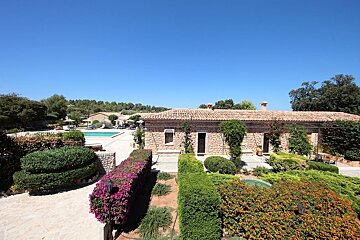 A sunny view of stone buildings with red tiled roofs, a blue swimming pool, and colorful, manicured gardens under a clear sky.