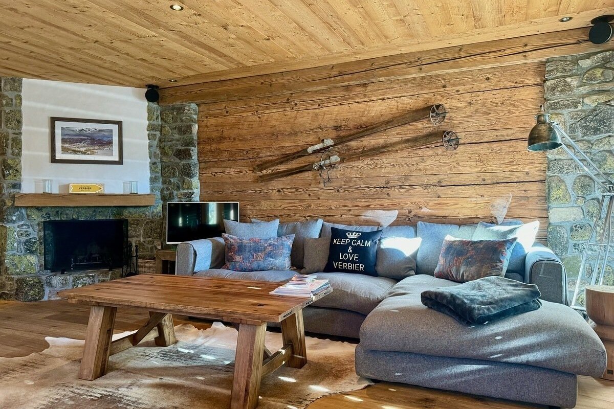 A cozy chalet living room with wood walls/ceiling, stone fireplace, large grey sofa, wooden coffee table, cowhide rug, and ski decor.