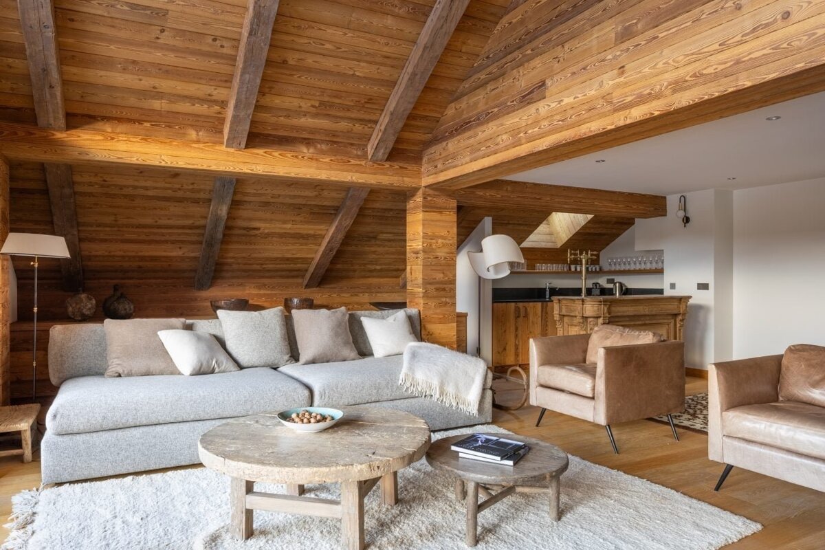 A cozy, rustic living room with extensive wood paneling, exposed beams, a large grey sofa, leather armchairs, and round wooden tables on a white rug.
