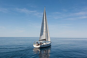A white sailboat with its large sails raised glides across calm, deep blue water under a clear sky.