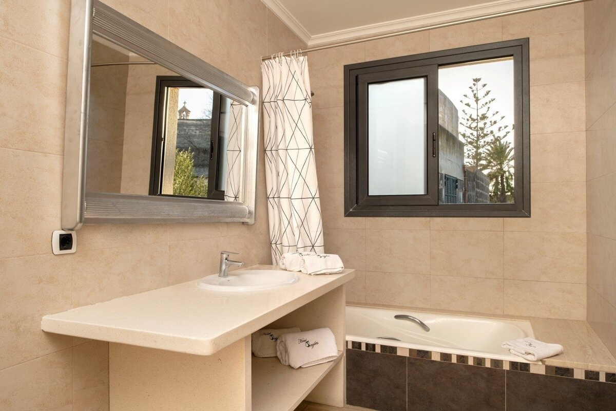 Light-toned tiled bathroom with a vanity, sink, large mirror, patterned shower curtain, window revealing trees, and a built-in bathtub.