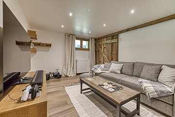 An inviting living room with a grey sectional, wooden coffee table (tic-tac-toe), TV, rustic wooden wall, and warm recessed lighting.