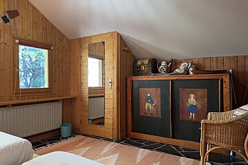 Cozy, wood-paneled attic room with a window, radiator, and a cabinet featuring folk art. A pink rug covers part of the stone floor.