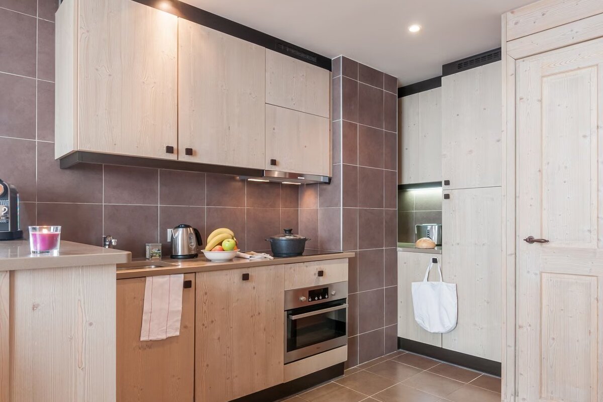 A modern kitchen with light wood cabinetry, brown tiled walls, stainless steel appliances, and a fruit bowl on the counter.