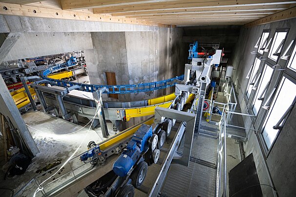 An intricate gondola or cable car station's machinery is housed indoors. Features include tracks, wheels, motors, concrete walls, a wooden ceiling, and a row of windows.