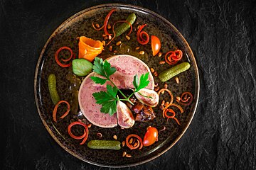 A gourmet slice of pรขtรฉ is elegantly presented on a dark plate, surrounded by gherkins, figs, parsley, colorful vegetable curls, and pine nuts.