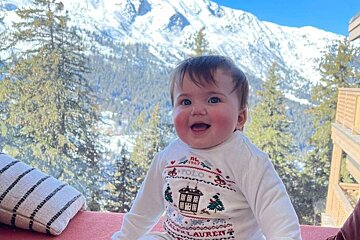 A happy baby in a holiday sweater smiles, with snowy mountains and evergreen trees behind them under a blue sky.