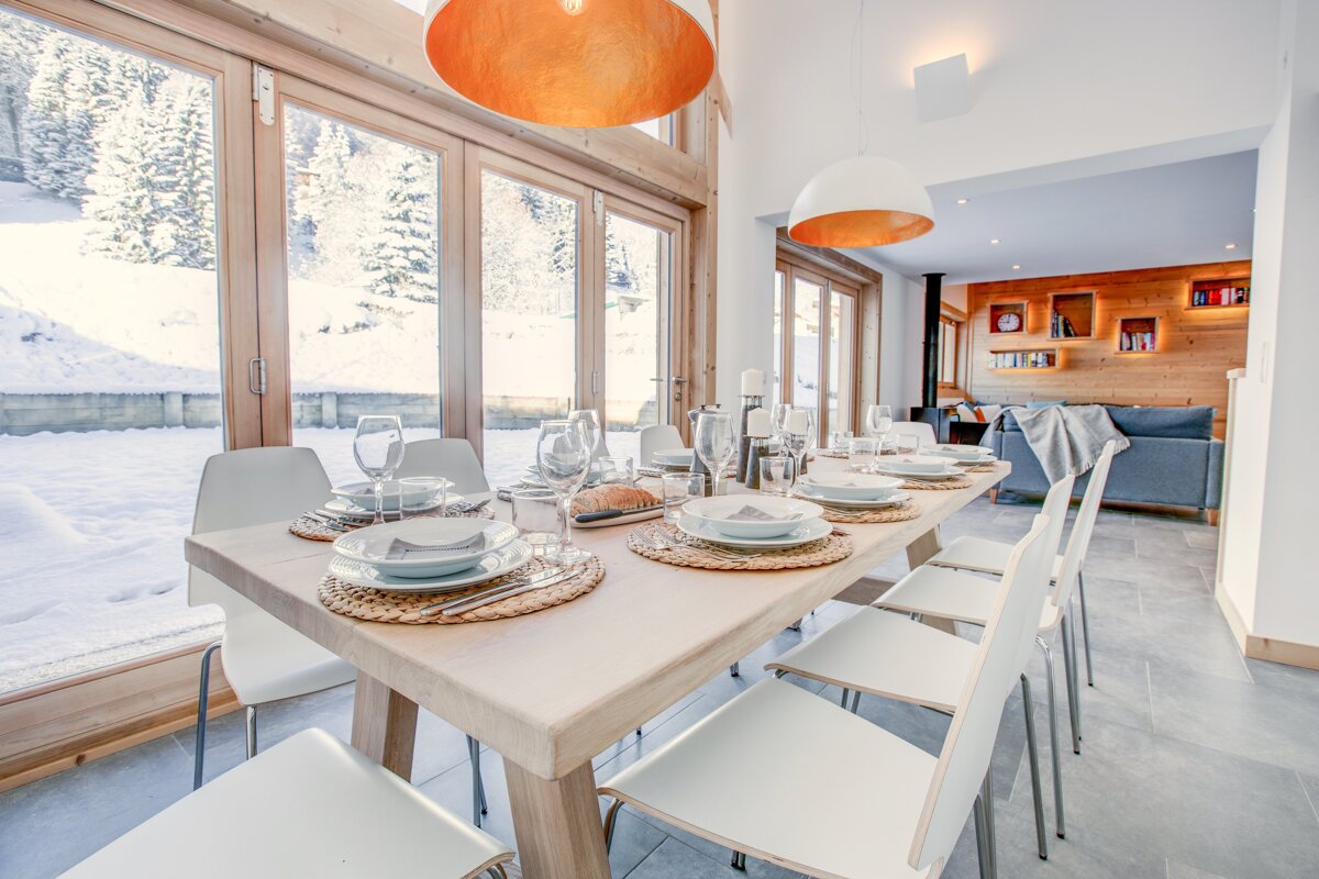 Bright modern dining room with a long table set for guests, featuring large windows with snowy winter views. Adjoining cozy living area.