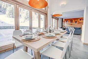 Bright modern dining room with a long table set for guests, featuring large windows with snowy winter views. Adjoining cozy living area.