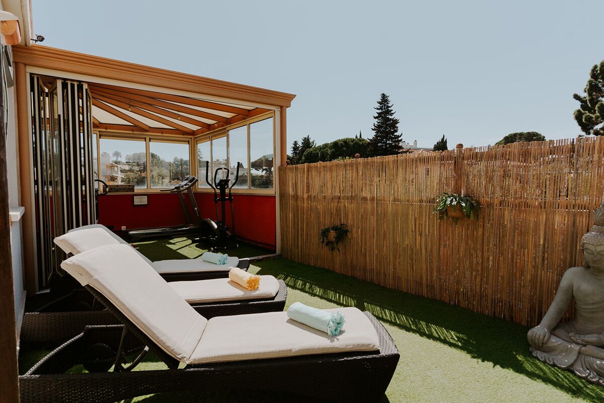 Sunny terrace with sun loungers, artificial grass, bamboo fence, and a Buddha statue. Adjacent glass-enclosed area has a treadmill and elliptical.