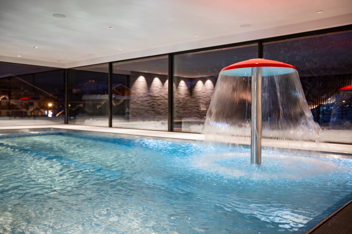 A modern indoor swimming pool with a red mushroom-shaped water cascade. Large windows offer views of a snow-covered evening landscape.