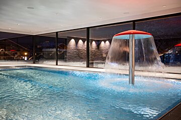 A modern indoor swimming pool with a red mushroom-shaped water cascade. Large windows offer views of a snow-covered evening landscape.