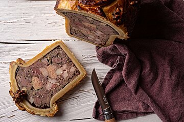 A savory pâté en croûte, sliced open to show its layered meat and fat filling, beside a cut piece, knife, and burgundy cloth on white wood.