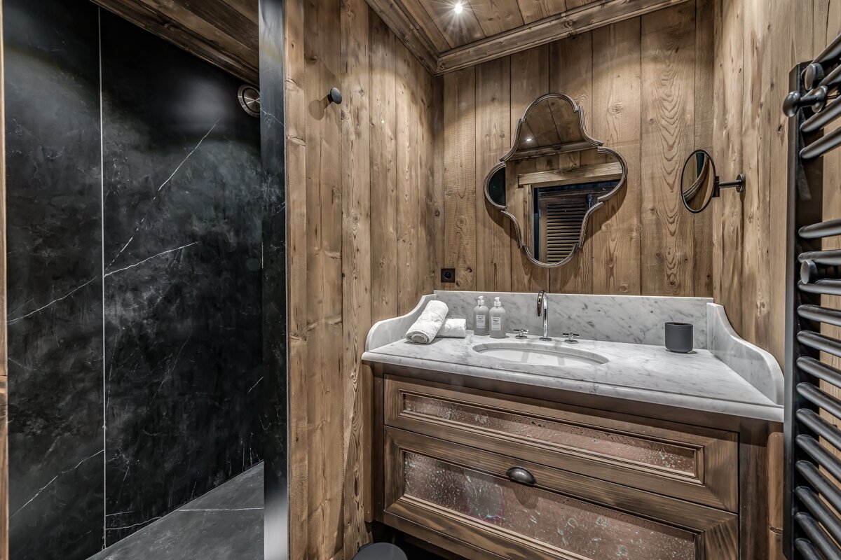A chalet-style bathroom featuring light wood paneled walls, a sleek black marble shower, and a stylish vanity with a white marble top and decorative mirror.