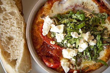 Baked eggs in tomato sauce, topped with feta, fresh herbs, and oil, served with a side of crusty bread.