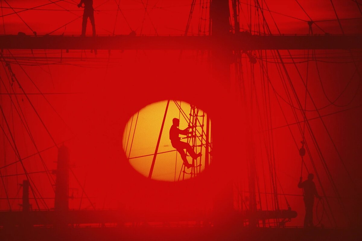 Fiery red image of a ship's rigging with silhouetted sailors. One climbs a ladder, framed perfectly by the glowing sun.