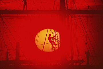 Fiery red image of a ship's rigging with silhouetted sailors. One climbs a ladder, framed perfectly by the glowing sun.