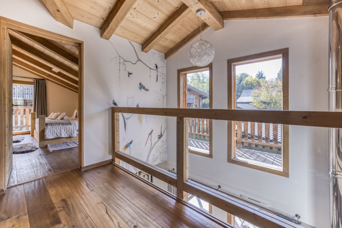 A bright, cozy chalet interior features exposed wooden beams and floors. A bird mural adorns a wall next to a glass and wood railing overlooking a lower level, with a bedroom visible through a doorway