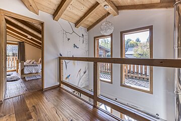 A bright, cozy chalet interior features exposed wooden beams and floors. A bird mural adorns a wall next to a glass and wood railing overlooking a lower level, with a bedroom visible through a doorway