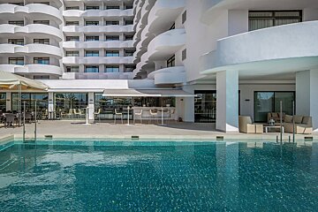 Modern white hotel with wavy balconies, a blue swimming pool, and sunny outdoor bar/lounge areas.