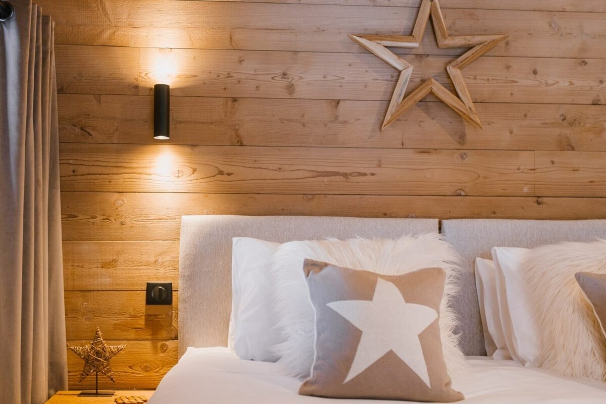A cozy, rustic bedroom featuring a wood-paneled wall with warm sconce lights, a plush bed with star pillows, and wooden star decor.