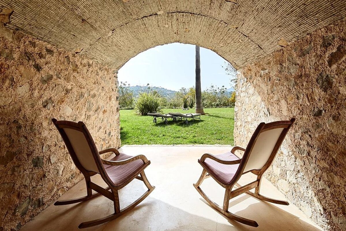 Two rocking chairs under a stone archway look out onto a lush green garden with lounge chairs and distant hills.
