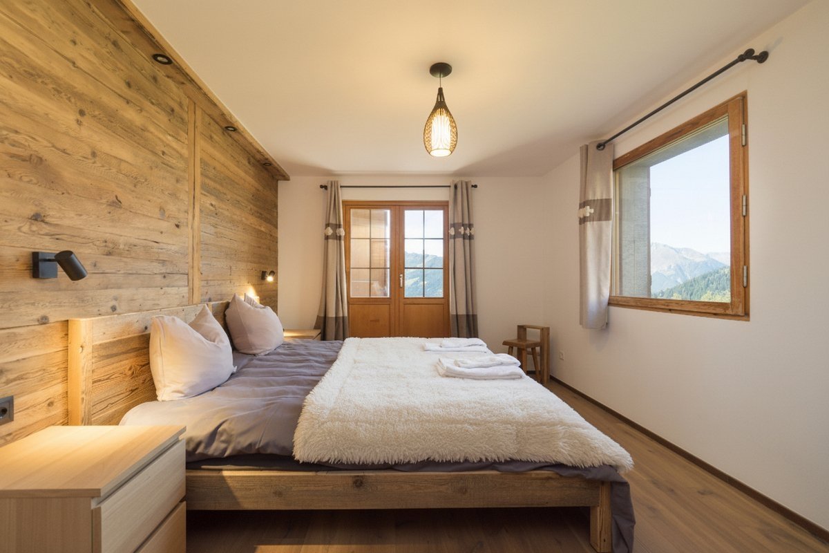 A modern bedroom featuring warm wood paneling, a large bed with fluffy blankets, and stunning mountain views from the windows and balcony doors.