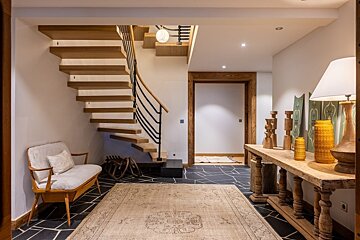 A stylish modern rustic foyer with a floating wooden staircase, black slate floor, rug, and a decorated wooden console table. A small bench sits nearby.