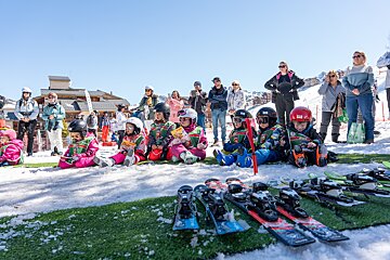 Young children in ski gear sit on snowy turf with adults watching, surrounded by skis and a sunny mountain resort backdrop.