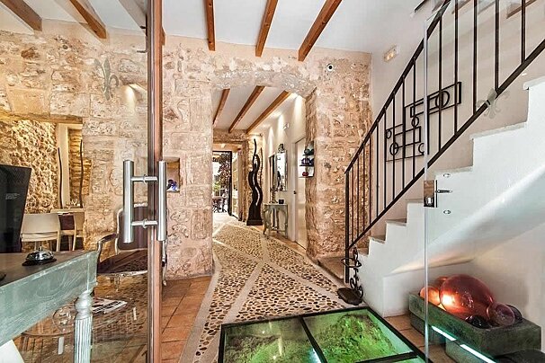 A rustic interior featuring stone walls, a pebbled path, wooden beams, a staircase, and glass details. An archway leads to a dining area.