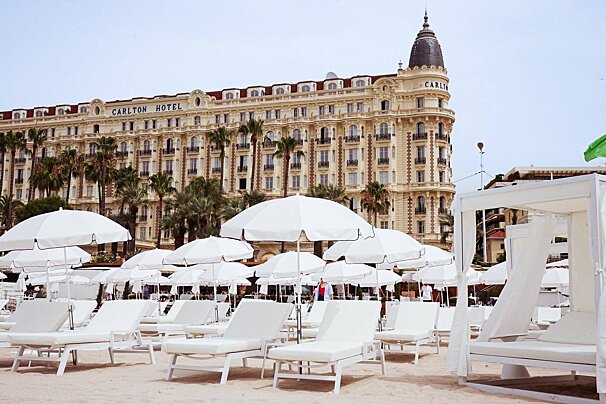 The grand Carlton Hotel overlooks a sandy beach lined with rows of white lounge chairs and umbrellas, backed by palm trees.