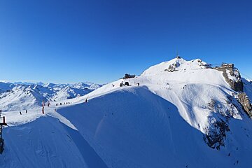 Vast snow-covered mountains under a clear blue sky, with ski slopes winding down and buildings visible on the peaks.