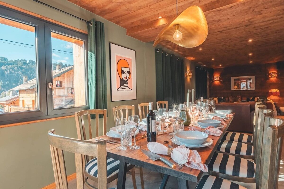 A stylish dining area with a rustic wooden table set for dinner, modern art, and a view of mountain homes from the window.