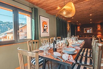 A stylish dining area with a rustic wooden table set for dinner, modern art, and a view of mountain homes from the window.