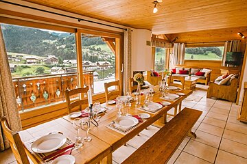 A warm, wood-paneled chalet interior features a long dining table set, large windows with a stunning mountain village view, and a cozy living area.