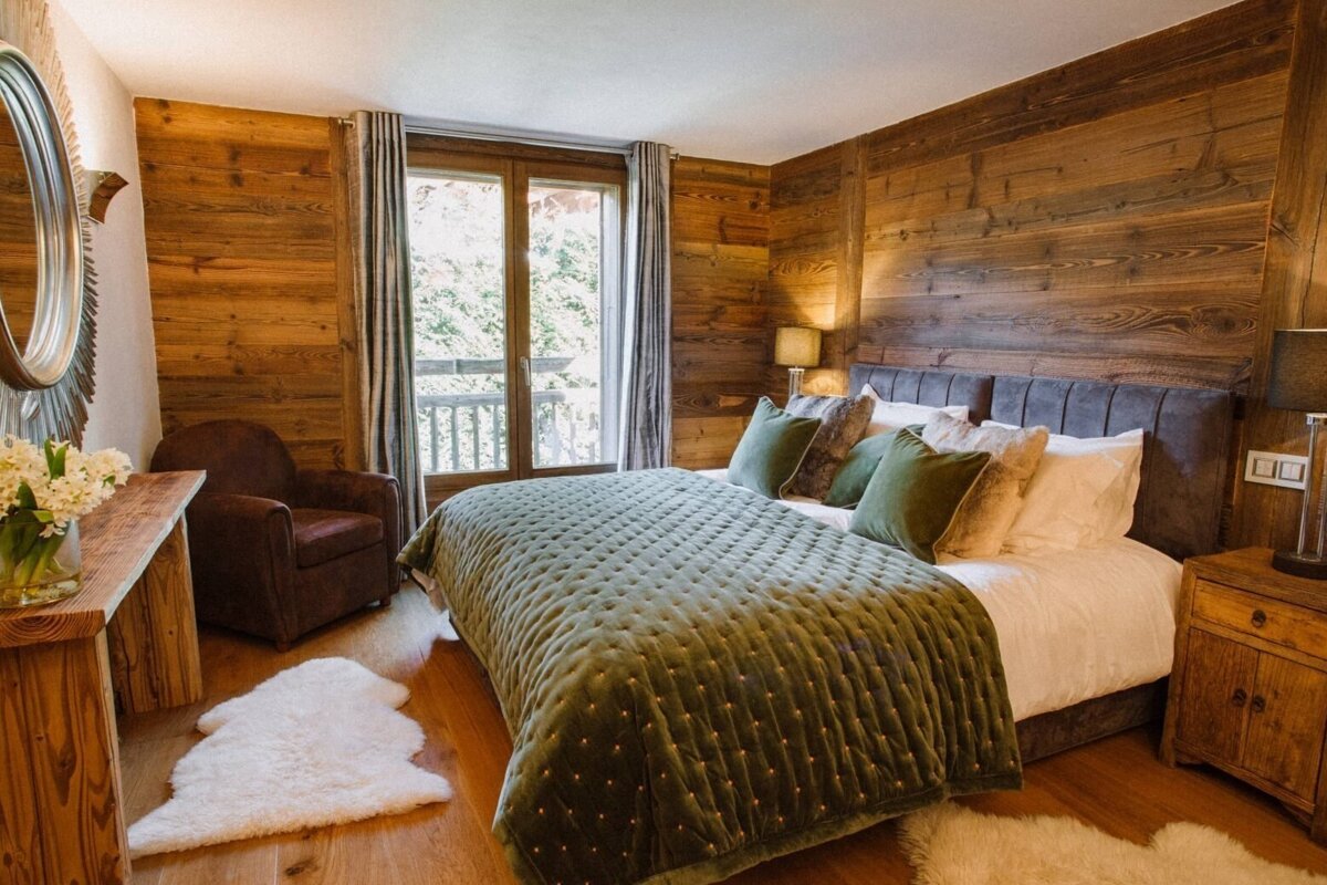 A cozy, chalet-style bedroom with wood-paneled walls, a luxurious green quilted bedspread, and a large window overlooking trees, offering a warm retreat.
