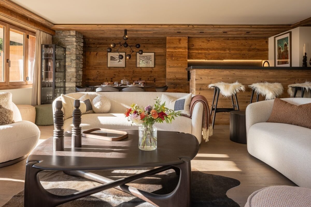 Chic chalet living room with warm wood walls, white sofas, elegant coffee table, and a bar area with fur stools. Bright and inviting.