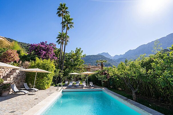 A sun-drenched outdoor swimming pool with lounge chairs, surrounded by lush tropical greenery, tall palm trees, and distant mountains.