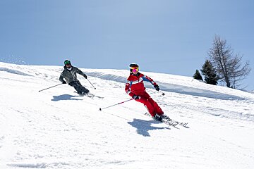Two skiers glide down a sunny, snow-covered mountain slope under a clear blue sky, carving fresh tracks.