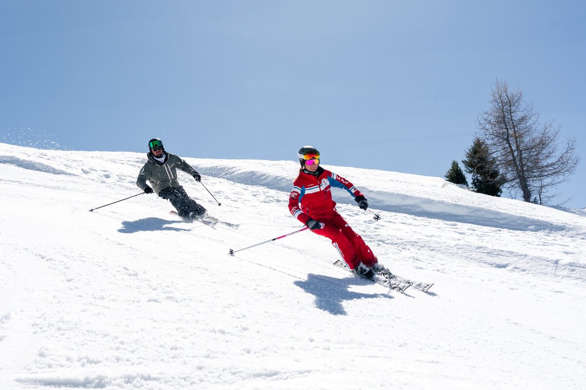 Two skiers carve down a sunny, snowy slope on a clear day, one in grey and one in red.