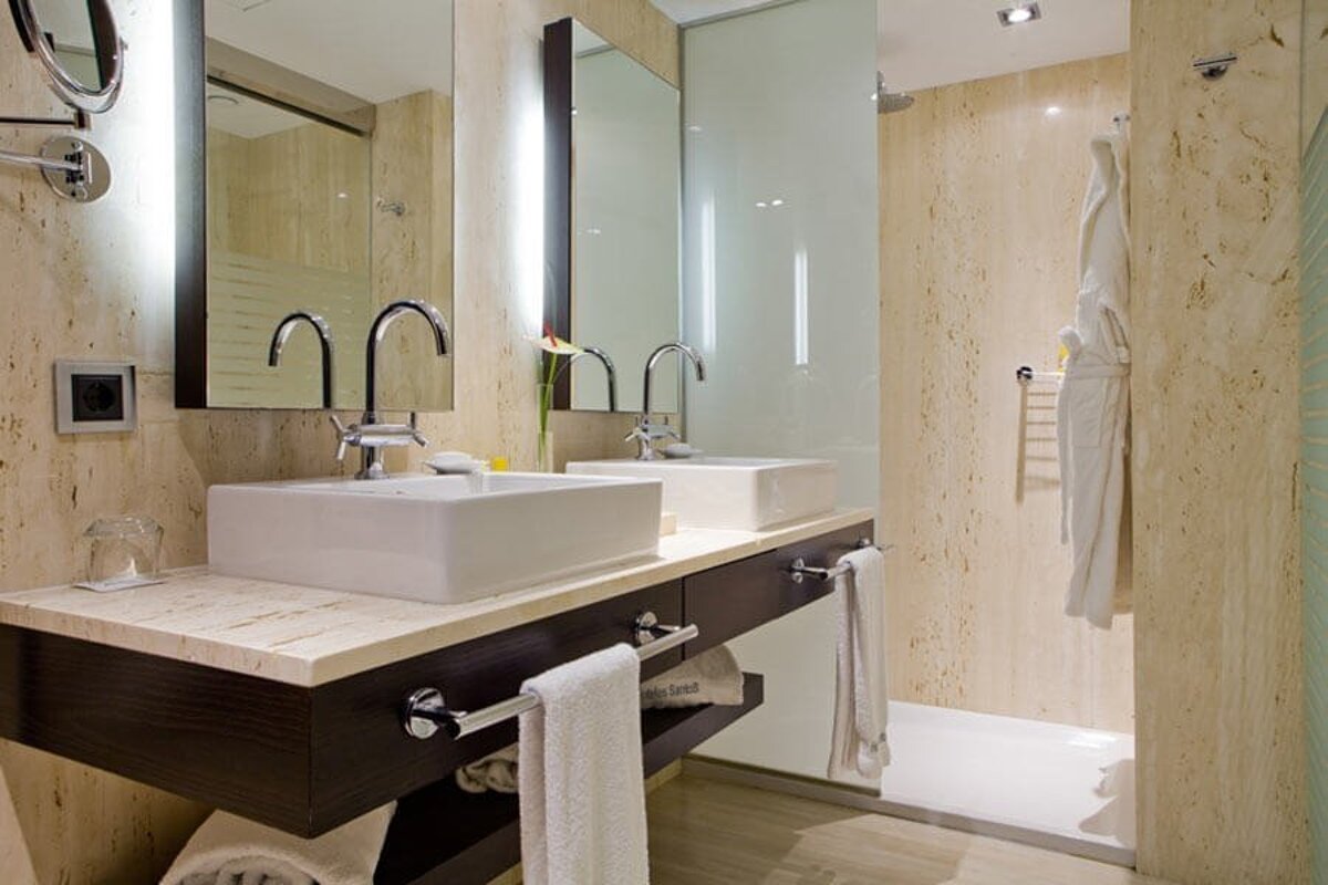 Modern bathroom with dual white vessel sinks on a marble vanity, large mirrors, and textured beige walls. A shower stall with a white bathrobe is on the right.