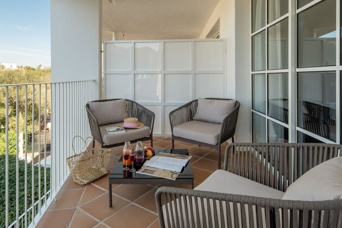 A sunny balcony with modern outdoor seating, a table with drinks and a magazine. Features a white privacy screen and a lush green view.