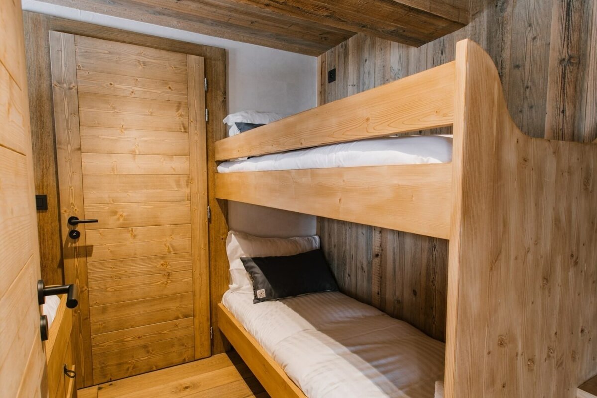 A rustic room features light wooden bunk beds with white bedding and dark pillows, set against plank wood walls, a wooden door, and a matching ceiling.