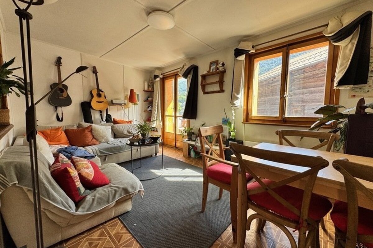 Cozy living and dining room with a sofa, two guitars on the wall, wooden dining table and chairs, and a window letting in sunlight.