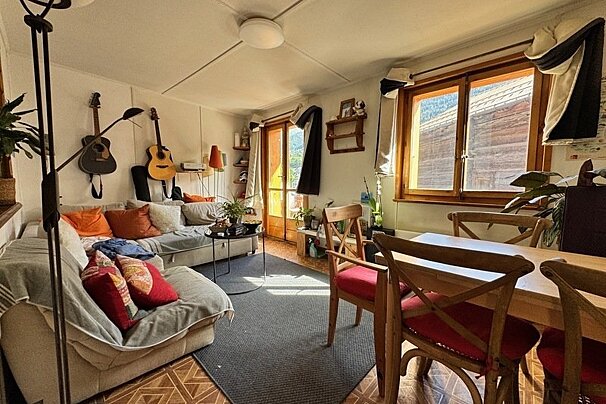 Cozy living and dining room with a sofa, two guitars on the wall, wooden dining table and chairs, and a window letting in sunlight.