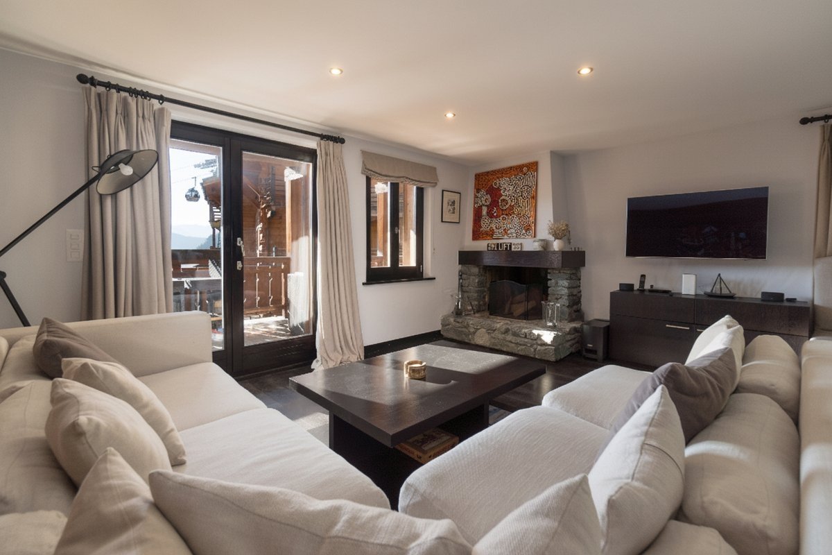 Inviting mountain chalet living room with comfy white sofas, a stone fireplace, TV, and a balcony overlooking a scenic mountainscape.