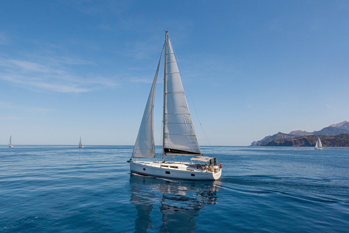 A white sailing yacht glides on calm blue water under a clear sky, with mountains in the background.
