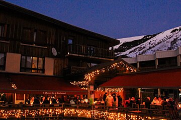 A warmly lit mountain restaurant patio bustling with diners under string lights at dusk. Snowy peaks loom in the background.